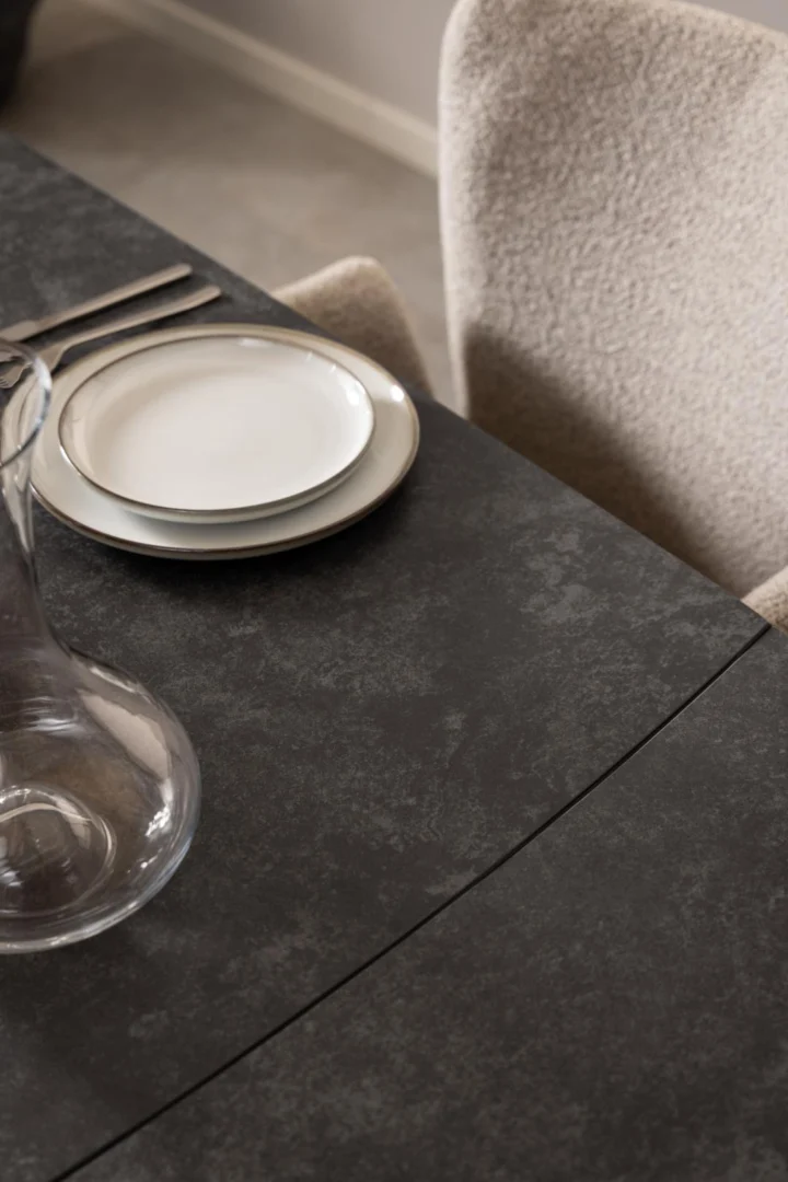 Een close-up van de Eettafel Nieuwe Sfeer (97 x 75 cm), met een rechthoekig glazen blad, een kan van helder glas, gestapelde witte borden, zilverwerk en stoelen met beige textuur op een donker, steenachtig oppervlak.