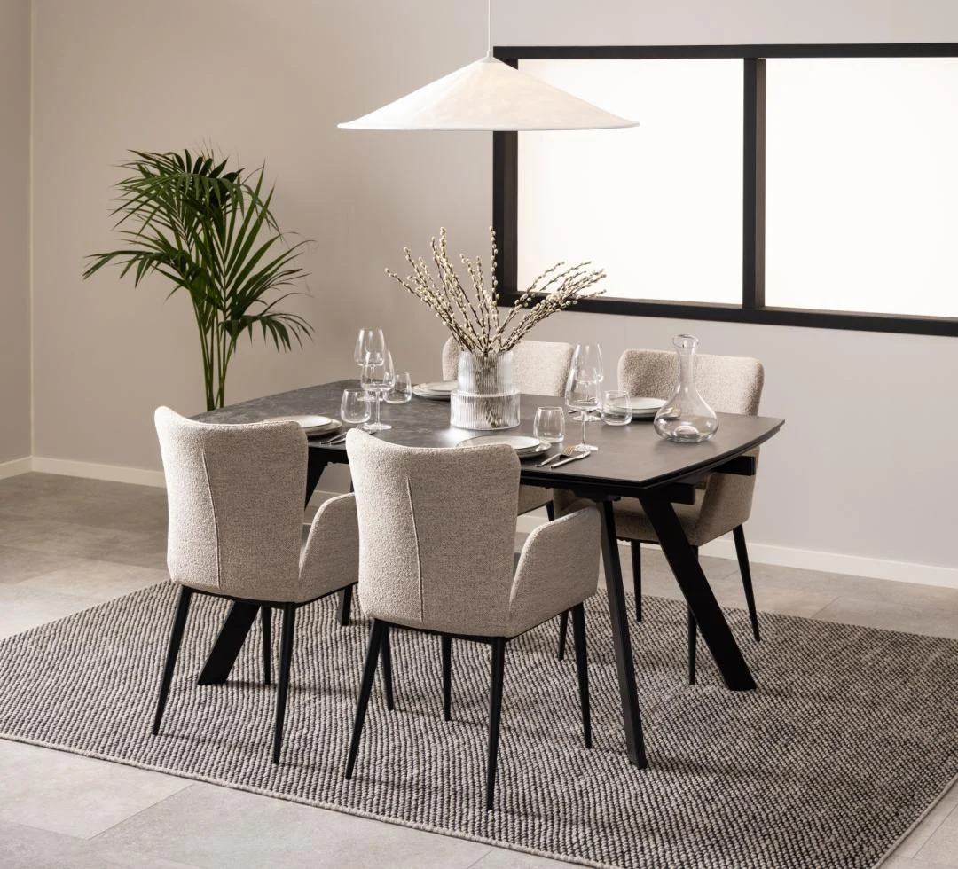 In een moderne eetkamer staat de Eettafel Nieuwe Sfeer 97 x 75 CM, een rechthoekige glazen tafel, met vier beige gestoffeerde stoelen, een grote hanglamp, een plant in pot in de hoek, een vloerkleed met structuur op een tegelvloer en een vaas met takken als decor.