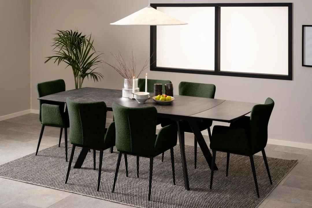 Een moderne eetkamer met de Eettafel Nieuwe Sfeer 97 x 75 CM rechthoek glas als middelpunt, omringd door zes donkergroene stoelen, vazen, kaarsen, een fruitschaal, hanglamp, hoekplant en een structuur vloerkleed eronder.