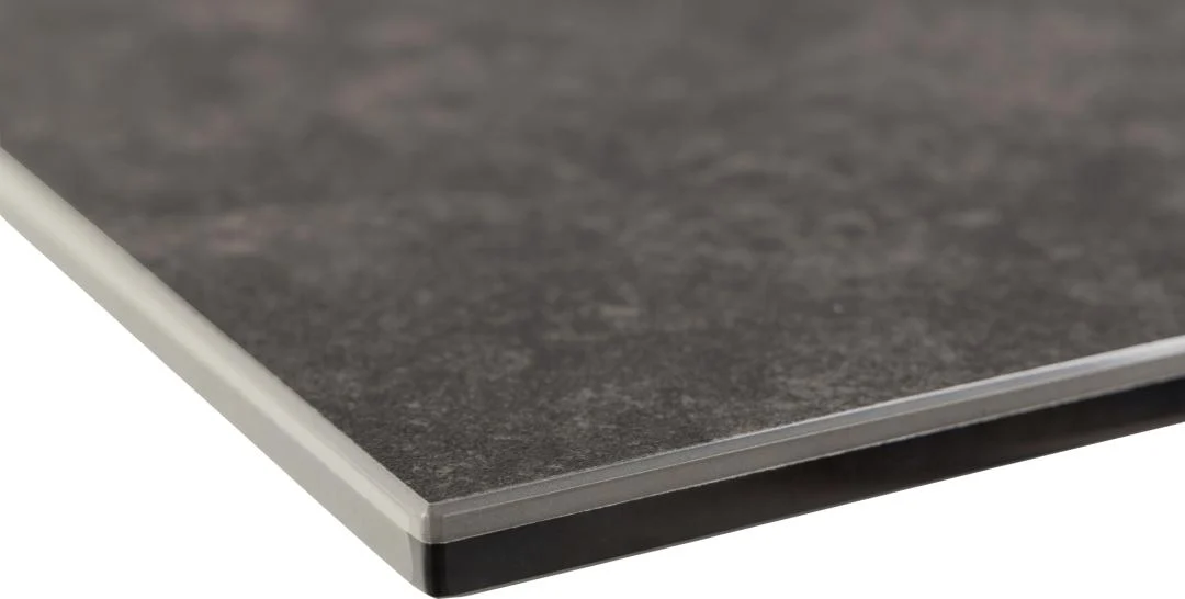 Close-up van de hoek van de Eettafel Irwine 90 x 76 CM Rechthoek, met een dun, rechthoekig donkergrijs keramiek blad met een gestructureerd oppervlak en gelaagde randdetails, tegen een witte achtergrond.