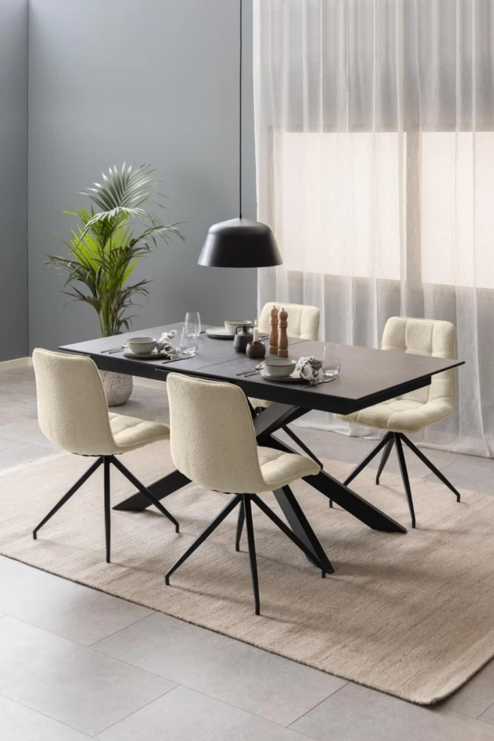 Een moderne eetkamer met de Eettafel Irwine 90 x 76 CM Rechthoek in zwart keramiek en staal, vier crèmekleurige stoelen met zwarte poten, servies, een zwarte hanglamp, een potplant en transparante gordijnen die zonlicht binnenlaten.