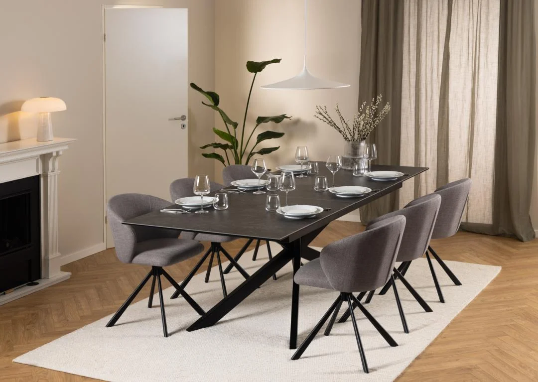 In een moderne eetkamer staat de Eettafel Nieuwe Sfeer 100 x 76 CM, een rechthoekige keramiek tafel met zes grijze stoelen met kussens. Borden, glazen en bestek zijn gedekt; neutrale muren, parketvloer, een vloerkleed, plant, transparante gordijnen en minimalistische inrichting maken de look compleet.