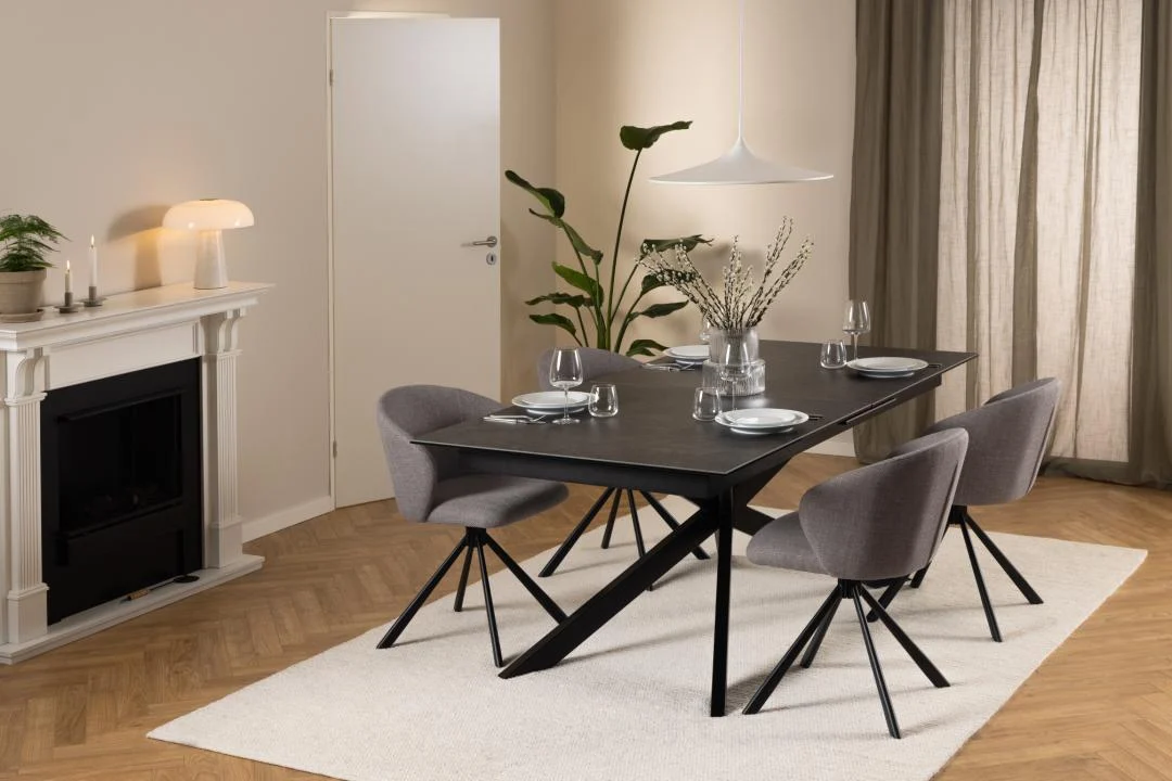 Een moderne eetkamer is voorzien van de Eettafel Nieuwe Sfeer 100 x 76 CM, een zwarte rechthoekige keramiek tafel voor vier personen met grijze stoelen, planten, een open haard en neutrale muren die worden verlicht door zacht natuurlijk licht via beige gordijnen.