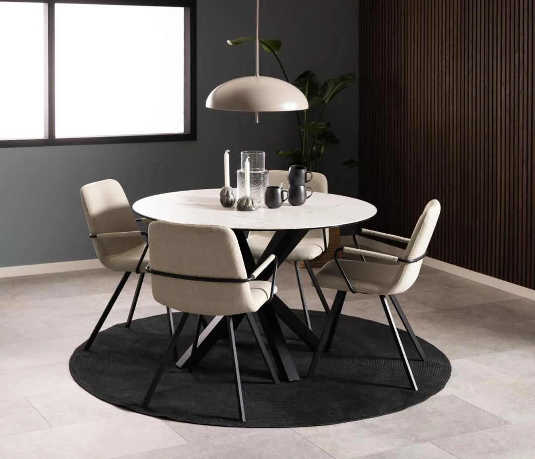 Een moderne eethoek met de Eettafel Hemel 119 CM Rond Keramiek Staal, vier beige stoelen met zwarte poten, een zwart rond vloerkleed, een hanglamp, decor voor het tafelblad, een plant in potten en een wand van donker houten panelen.