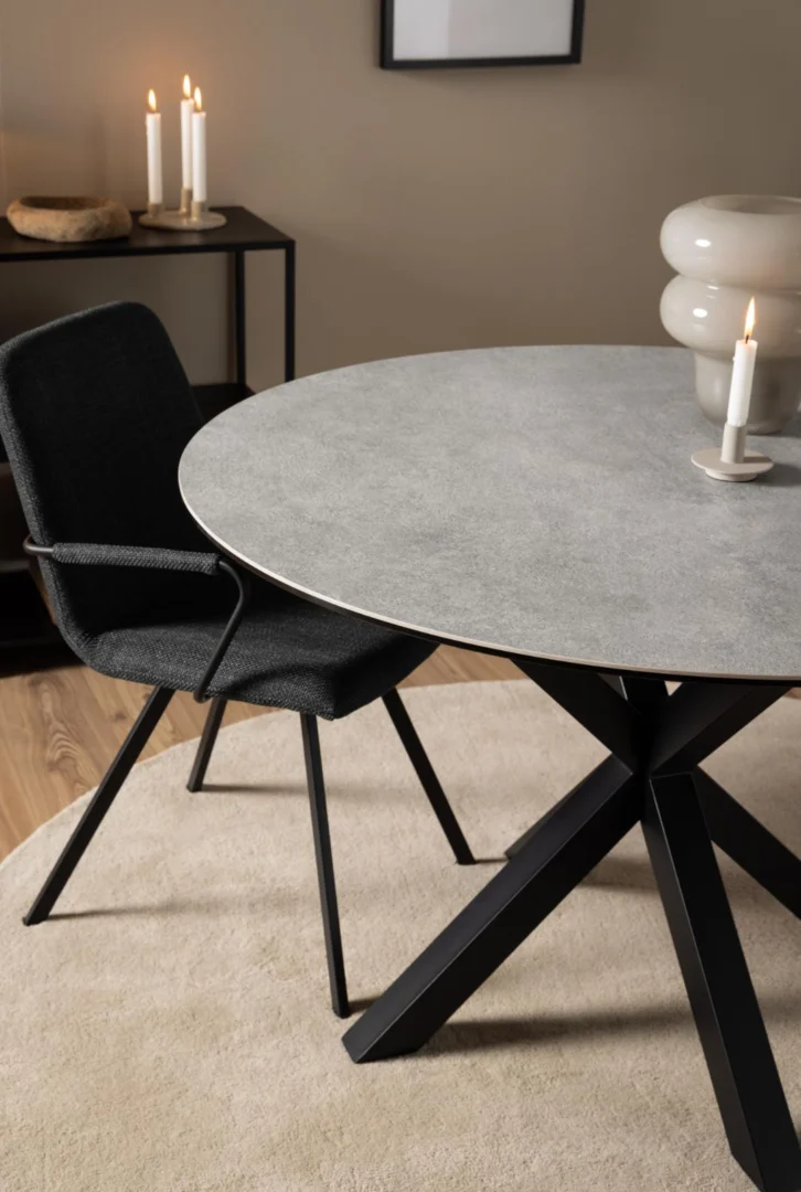 Een moderne eethoek met de Eettafel Nieuwe Sfeer 119 CM Rond Staal met een mat zwart onderstel, gecombineerd met een zwarte stoel en een gloeiende witte kaars. Meer kaarsen en decor staan op een zwarte console tegen een beige muur op de achtergrond.