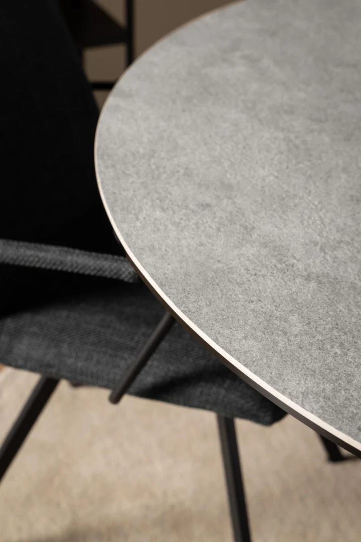 Een close-up van de Eettafel Nieuwe Sfeer 119 CM Rond Staal met een grijs stenen blad en mat zwart onderstel, in combinatie met een zwarte stoffen stoel op een lichtgekleurde vloer.
