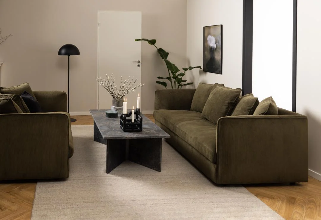 Een moderne woonkamer met olijfgroene banken, een Salontafel Bruin 70 x 42 CM Rechthoek Marmer, kaarsen, een vaas, beige vloerkleed, staande lamp, potplant en minimalistische muurkunst tegen neutrale muren en een houten vloer.