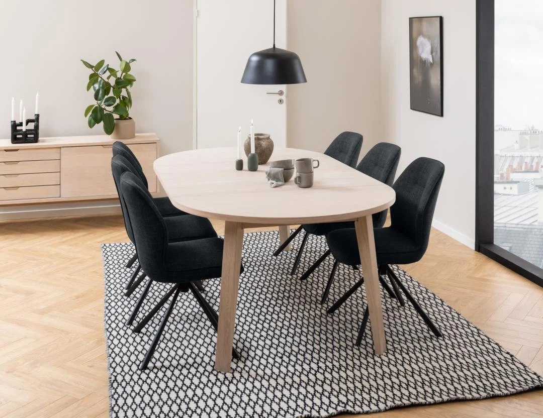 Een moderne eetkamer met de Eettafel 120 CM Rond Spaanplaat, zes zwarte stoelen met kussens, een zwarte hanglamp, geometrisch vloerkleed, dressoir met decor, plant in pot en ingelijste kunst bij het raam.