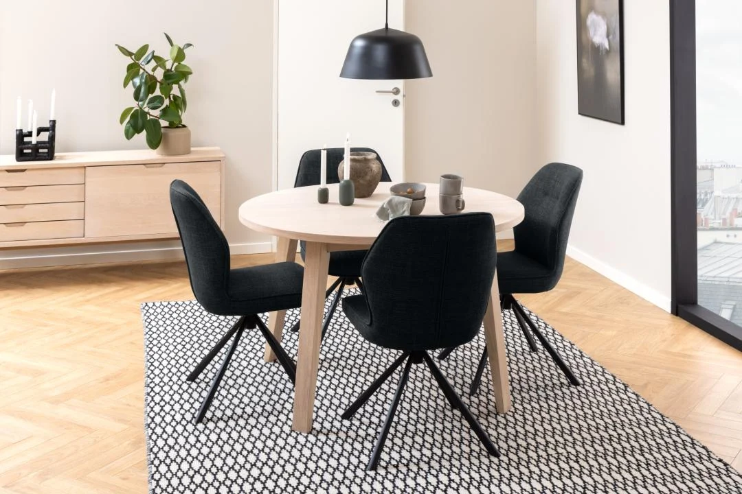 In een moderne eetkamer staan de Eettafel 120 CM Rond Spaanplaat, vier zwart gestoffeerde stoelen, een zwarte hanglamp en servies. Een dressoir met planten en decor staat tegen een witte muur op een vloerkleed met patroon.