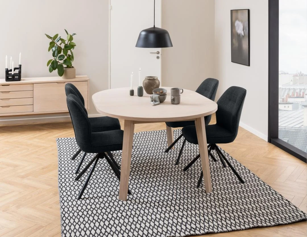 Een moderne eetkamer met de Eettafel 120 CM Rond Spaanplaat, vier donker gestoffeerde stoelen, een zwarte hanglamp, minimalistische inrichting, een dressoir met een plant en kaarsen en een geometrisch vloerkleed. Grote ramen laten de daken van de stad zien.