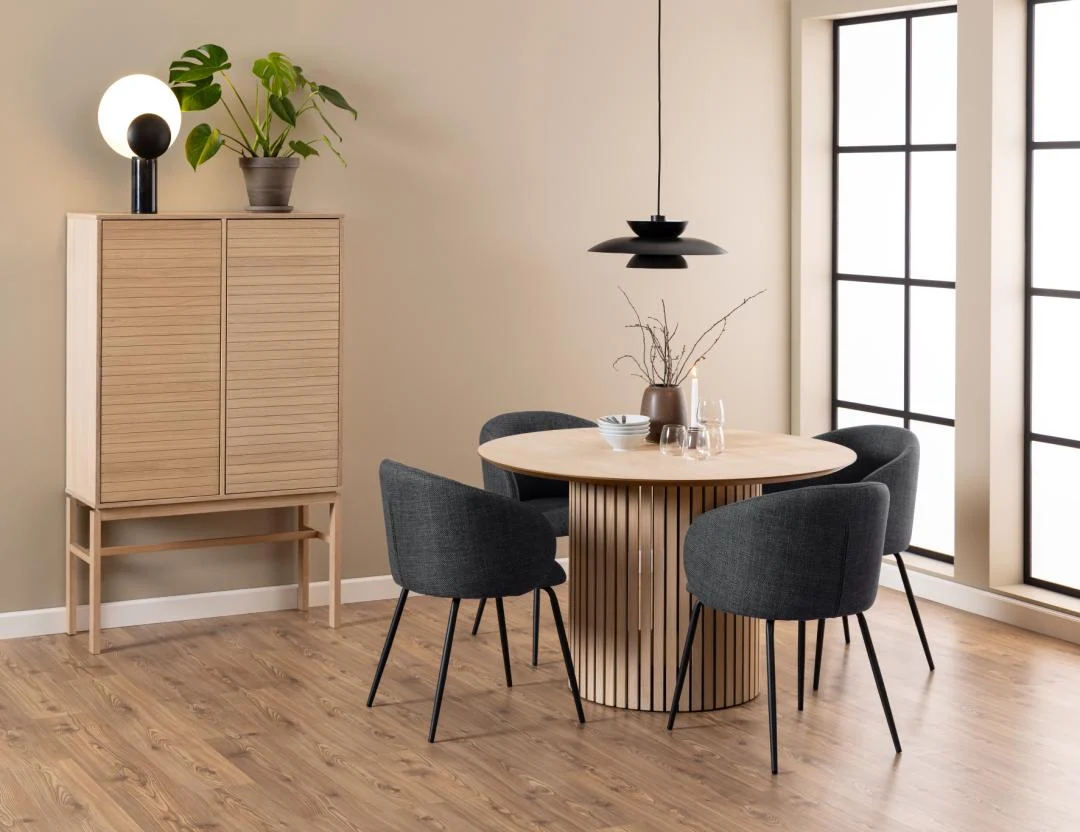 Een moderne eethoek met de Eettafel Nieuwe Sfeer 120 CM Rond (eikenhouten ronde tafel), vier donkergrijs gestoffeerde stoelen, een houten kast met daarop planten en een lamp, en grote ramen in rasterstijl die natuurlijk licht binnenbrengen.