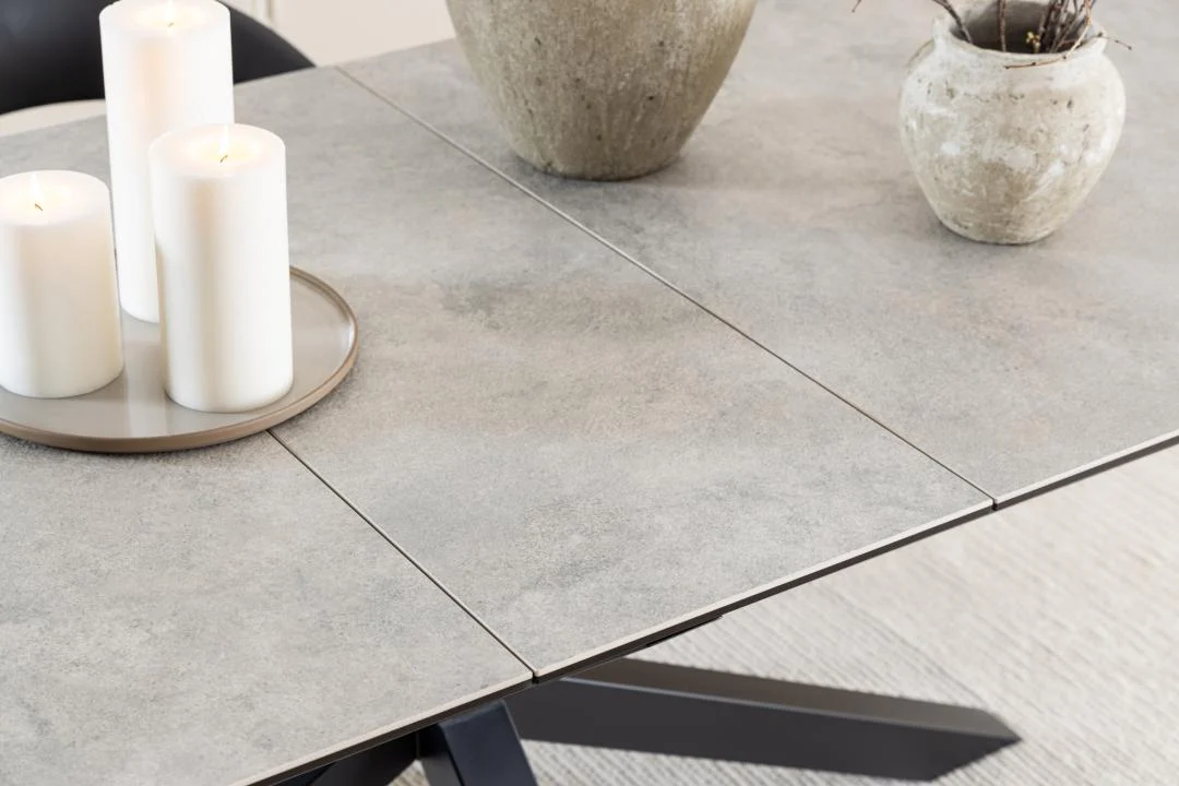 De Eettafel Hemel 100 x 76 CM Rechthoek, gemaakt van grijs keramiek en zwarte stalen poten, is gestyled met drie witte pilaarkaarsen op een dienblad en twee beige structuurvazen, geplaatst op een licht geweven vloerkleed.