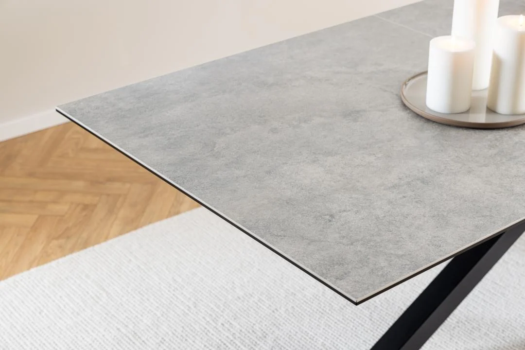 Een close-up van de Eettafel Hemel 100 x 76 CM Rechthoek, met een dun, steenachtig keramiek blad en zwarte stalen hoekpoten, geplaatst op een beige vloerkleed met een rond dienblad met witte pilaarkaarsen en een houten vloer ernaast.