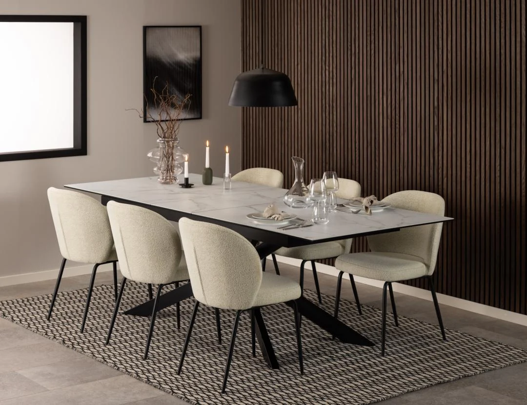 In een moderne eetkamer staan de Eettafel Irwine 240 x 100 CM Rechthoek Keramiek tafel, zes crèmekleurige stoelen, een zwarte hanglamp, kaarsen, glaswerk en een minimalistische inrichting op een gedessineerd tapijt met houten lambrisering en lichte muren.