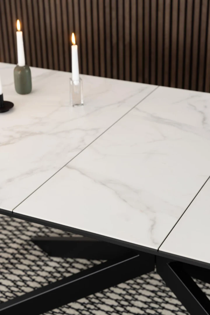De Eettafel Irwine 240 x 100 CM Rechthoek, met een keramisch marmeren blad en zwarte poten, is vormgegeven met brandende witte kaarsen in minimalistische houders. Hij staat op een gedessineerd vloerkleed met een houten lattenwand als achtergrond.