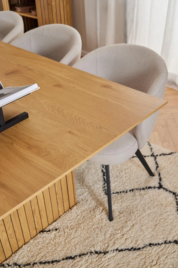 De Eettafel Nieuwe Sfeer 100 x 75 CM, een lichthouten rechthoekige MDF tafel, wordt gecombineerd met een beige gestoffeerde stoel op zwarte poten op een crèmekleurig vloerkleed met een eenvoudig zwart geometrisch patroon. Zacht licht valt door witte gordijnen op de achtergrond.