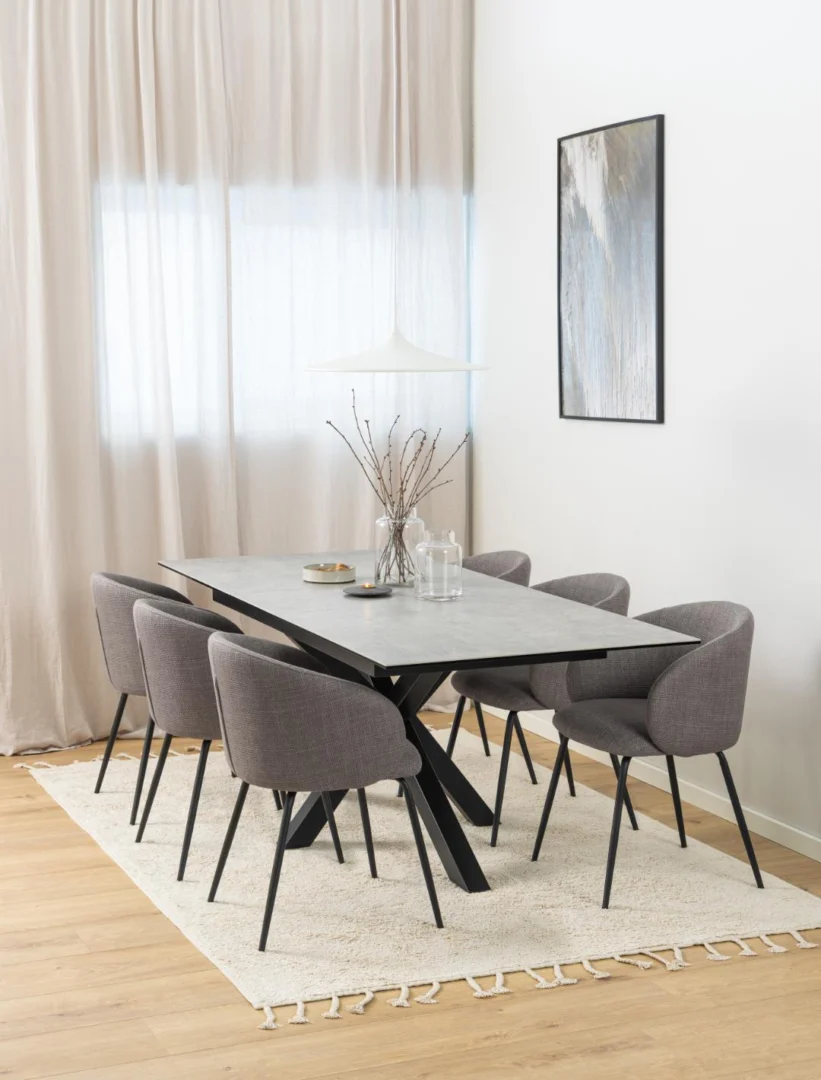 Een moderne eetkamer is voorzien van de Eettafel Nieuwe Sfeer 90 x 76 CM, een rechthoekige robuuste glazen tafel, zes grijze stoelen, een wit vloerkleed op lichthouten vloeren, beige gordijnen, minimalistische inrichting, hanglamp en abstracte muurkunst voor een elegante neutrale look.