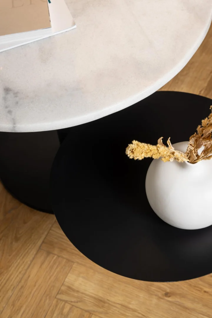 De Salontafel Zwart, Wit 45 CM Rond, een ronde wit met zwarte kunststof nestelset, heeft een gedeeltelijk overlappend ontwerp met een witte vaas met gedroogde gele bloemen, geplaatst op een houten vloer met visgraatmotief.