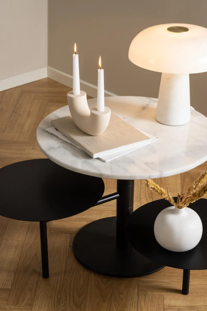 De Salontafel Zwart, Wit 45 CM Rond uit de Kunststof Nesting Set toont een open boek, een moderne witte kandelaar met twee brandende kaarsen en een witte lamp. Een kleine witte vaas met gedroogde bloemen rust op de onderste zwarte plank boven de parketvloer.