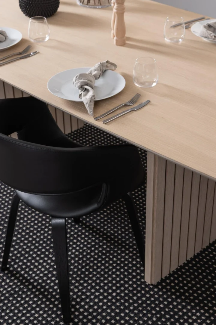 De Eettafel Nieuwe Sfeer 105 x 75 CM, een robuuste rechthoekige MDF tafel, is gecombineerd met witte borden, grijze servetten, glaswerk, bestek en een zwarte stoel op een zwart-wit gedessineerd vloerkleed.