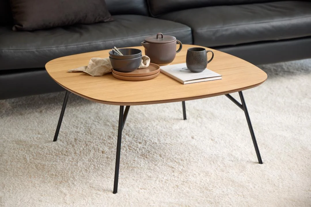 De Salontafel Zwart 90 x 90 CM Rond, gemaakt van eikenfineer, bevat een theepot, kop, kom met keukengerei, servet en boek. Hij staat op een licht tapijt voor een zwartleren bank met donkere kussens.