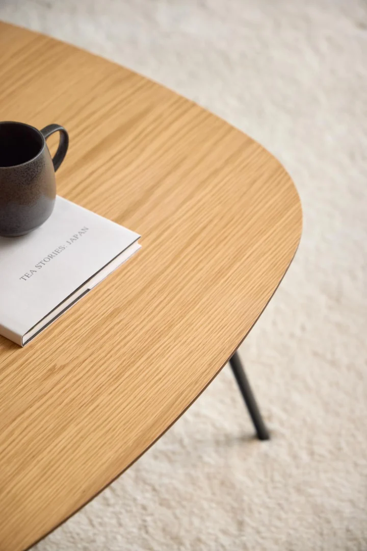 Een close-up van de Salontafel Zwart 90 x 90 CM Rond in eikenfineer, met een zwarte keramische mok en een gesloten wit boek op het oppervlak, tegen een lichtgekleurde getextureerde tapijtachtergrond.