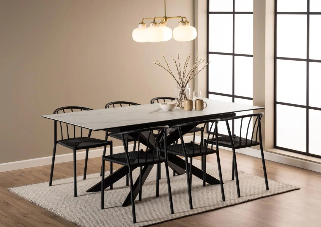 Een moderne eetkamer is voorzien van de Eettafel Nieuwe Sfeer 90 x 76 CM, een rechthoekige tafel van keramiek met zes zwarte stoelen, een beige vloerkleed, een gouden globe kroonluchter, een vaas met gedroogde takken, mokken, borden en grote ramen voor natuurlijk licht.