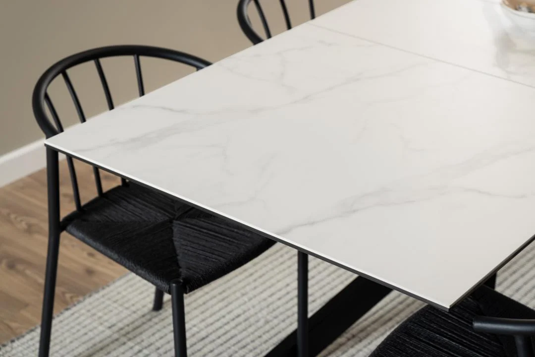 Een close-up van de Eettafel Nieuwe Sfeer 90 x 76 CM, een moderne rechthoekige eettafel van keramiek met een blad in wit marmerpatroon, gekoppeld aan zwarte stoelen met geweven zitting op een licht getextureerd tapijt.