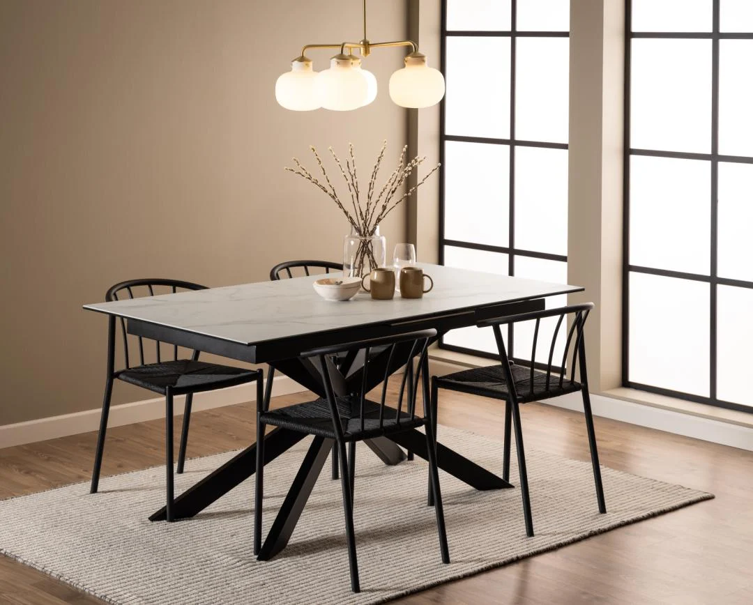 In een moderne eetkamer staan de Eettafel Nieuwe Sfeer (90 x 76 cm) rechthoek keramiek tafel, vier zwarte stoelen, een beige vloerkleed en een middelpunt van takken. Daarboven hangen drie globe hangers met grote ramen die natuurlijk licht binnenbrengen.