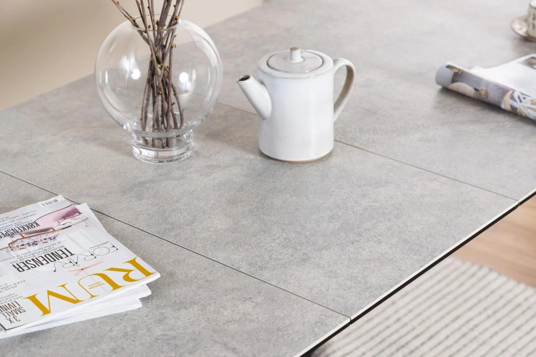 Een witte theepot, een vaas van helder glas met takken, een opgerold tijdschrift en gestapelde tijdschriften liggen op de lichtgrijze Eettafel Irwine 90 x 76 CM Rechthoek (keramiek, staal). Een beige en wit gestreept vloerkleed is gedeeltelijk zichtbaar aan de onderkant.