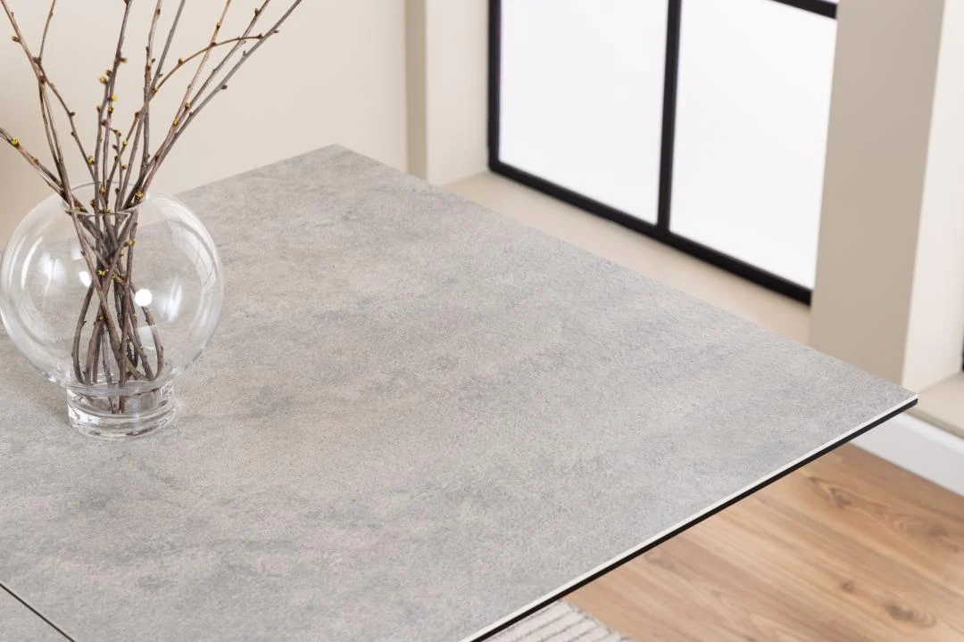 Een ronde glazen vaas met dunne, kale takken staat op de hoek van de Eettafel Irwine 90 x 76 CM Rechthoek, een tafel van keramiek en staal, naast een raam en een houten vloer.