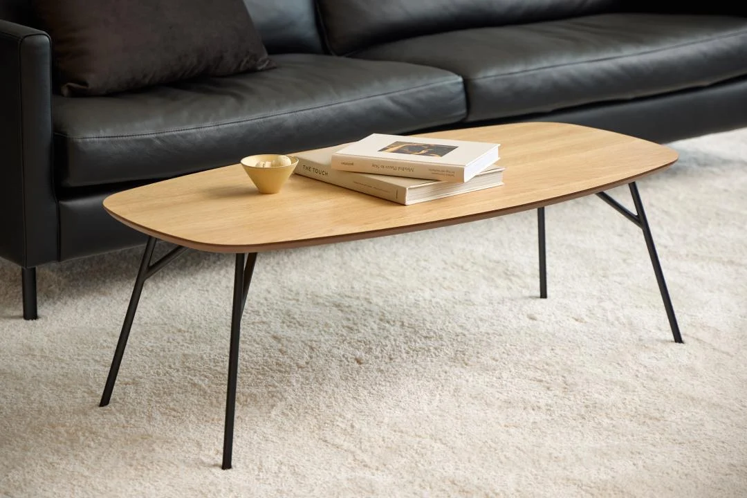 De Salontafel Zwart 40 CM Rond Eikenfineer ligt op een lichtgekleurd tapijt voor een zwartleren bank, met twee gestapelde boeken en een kleine beige schaal op het oppervlak.