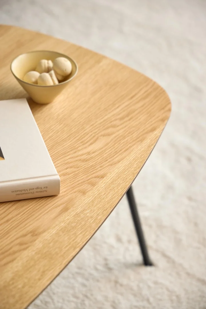 Een close-up van de Salontafel Zwart 40 CM Rond Eikenfineer met een beige decorschaal en een gesloten boek op het houten oppervlak; op de achtergrond verschijnt een licht, gestructureerd tapijt.