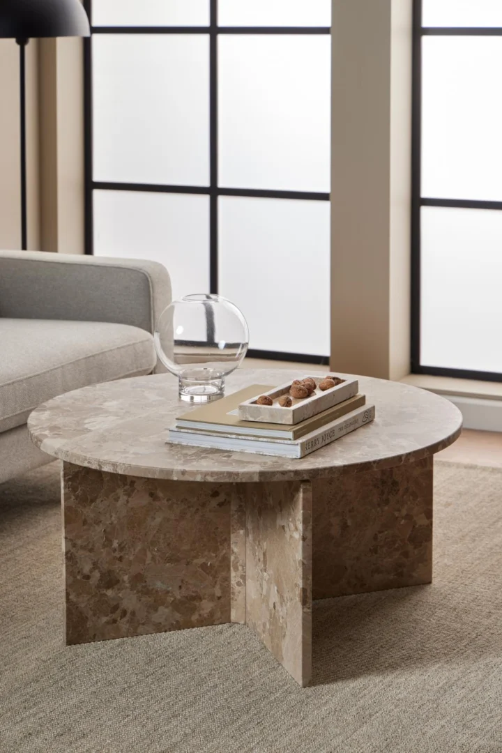 De Salontafel Bruin, Roest 90 CM Rond Marmer, met een uniek kruisvormig onderstel, houdt een dienblad met boeken en decor naast een lichtgrijze bank in een moderne woonkamer met grote rasterramen.