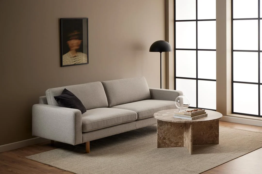 Een moderne woonkamer met een lichtgrijze bank, een zwarte staande lamp, de Salontafel Bruin, Roest 90 CM Rond Marmer met daarop boeken en een glazen beker, een beige vloerkleed, hoge ramen met zwarte lijst en een vaag portret aan de muur.