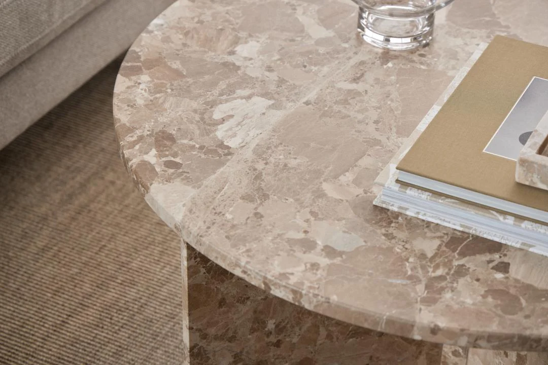 Een close-up van de Salontafel Bruin, Roest 90 CM Rond Marmer met een paar boeken en een glazen object erop, zittend op een vloerkleed met structuur naast een lichtgekleurde bank.