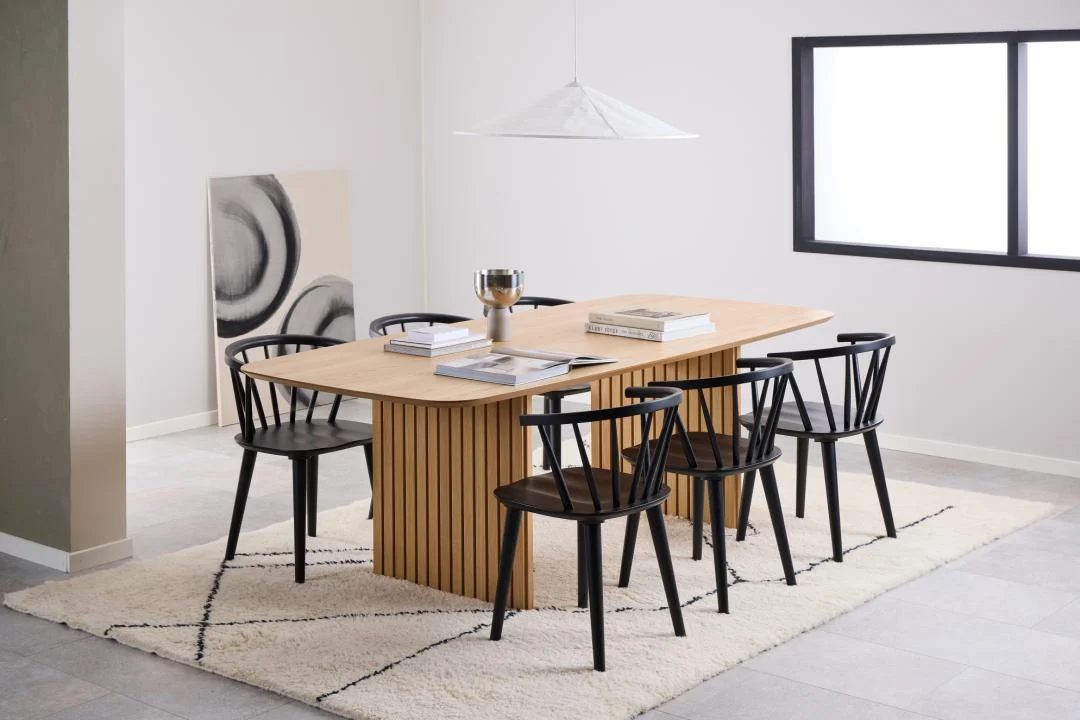In een moderne eetkamer staan de Eettafel Nieuwe Sfeer (105 x 75 cm), een rechthoekige eikenhouten tafel, zes zwarte stoelen, boeken en een schaal in het midden. De tafel rust op een licht geometrisch tapijt; abstracte kunst leunt bij een groot raam.