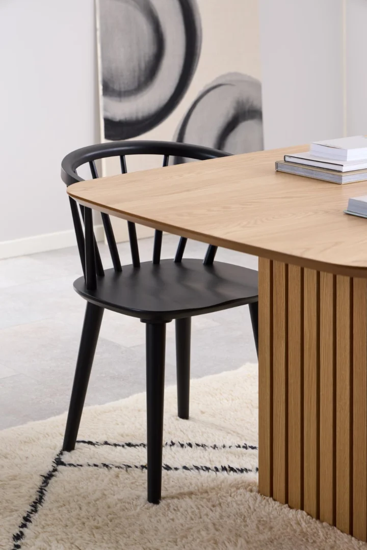 Een moderne zwarte houten stoel staat naast de Eettafel Nieuwe Sfeer 105 x 75 CM, een rechthoekige eiken tafel, op een wit vloerkleed met zwarte lijnen. Op de achtergrond is een abstract zwart-wit kunstwerk te zien.