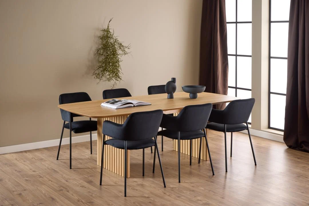 Een moderne eetkamer met de Eettafel Nieuwe Sfeer (105 x 75 cm) in rechthoekig eikenhout, zes zwarte stoelen, een decoratieve schaal, vazen, een open boek, grote ramen met bruine gordijnen, een lichthouten vloer en een groene muurplant.