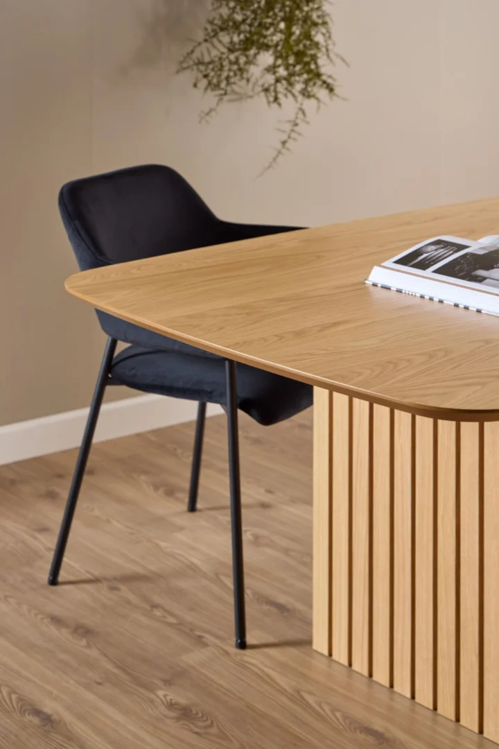 De Eettafel Nieuwe Sfeer 105 x 75 CM, een rechthoekige eiken tafel met verticale groeven, staat op een lichte houten vloer. Ernaast staat een zwarte stoel met kussens en slanke metalen poten en op het tafelblad ligt een open tijdschrift.