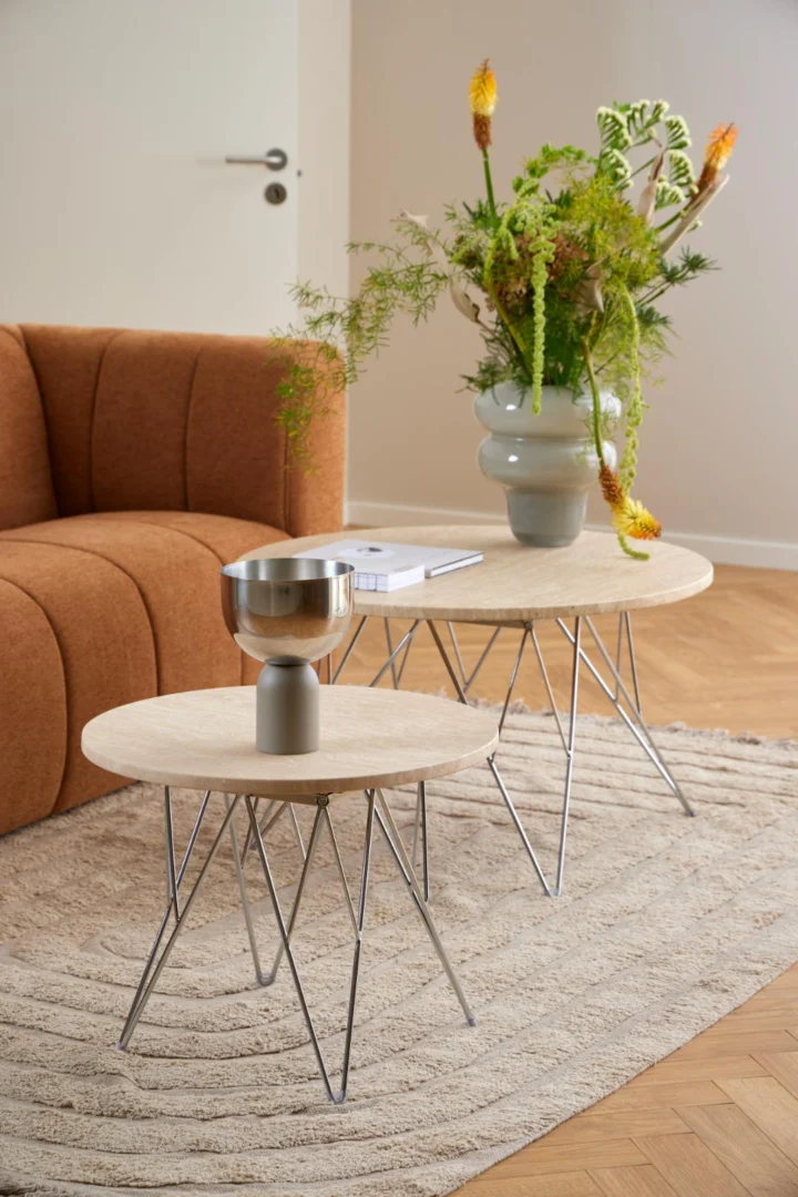 Een Salontafel Beige 80 CM Rond Kunststof ligt op een beige vloerkleed, gestileerd met een zilveren decoratieve schaal en een moderne vaas met daarin diverse soorten groen en bloemen. Op de achtergrond is een bruine bank te zien.