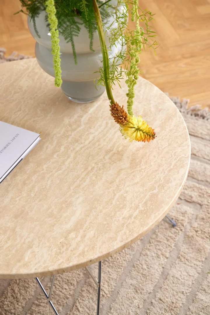 Een Salontafel Beige 80 CM Rond Kunststof met metalen poten toont een witte vaas met groen gebladerte en een oranje-gele bloem. Een deel van een open boek en een vloerkleed met structuur zijn zichtbaar op de houten vloer.