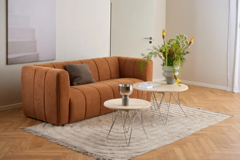 Een moderne woonkamer met een roestkleurige bank, bruin kussen, de Salontafel Beige 80 CM Rond Kunststof salontafel, grijs decoratief kopje, vaas met groene en gele bloemen op een beige vloerkleed op een houten vloer.