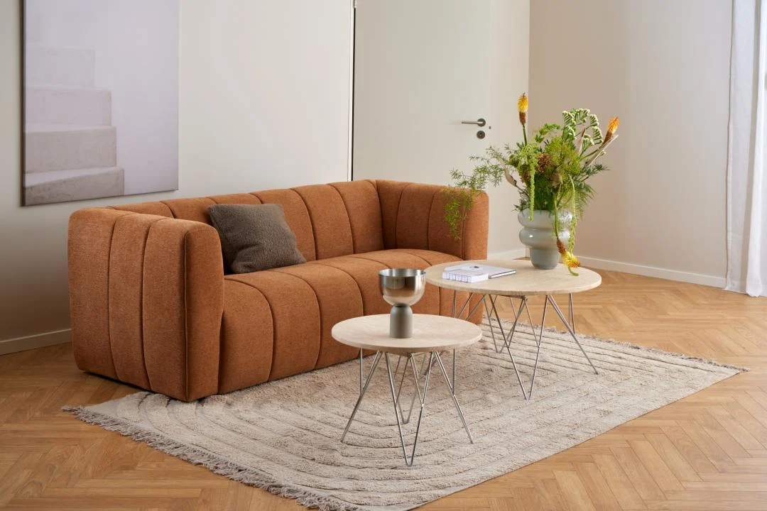 Een moderne woonkamer met een roestkleurige bank, bruin kussen, de Salontafel Beige 80 CM Rond Kunststof salontafel, grijs decoratief kopje, vaas met groene en gele bloemen op een beige vloerkleed op een houten vloer.