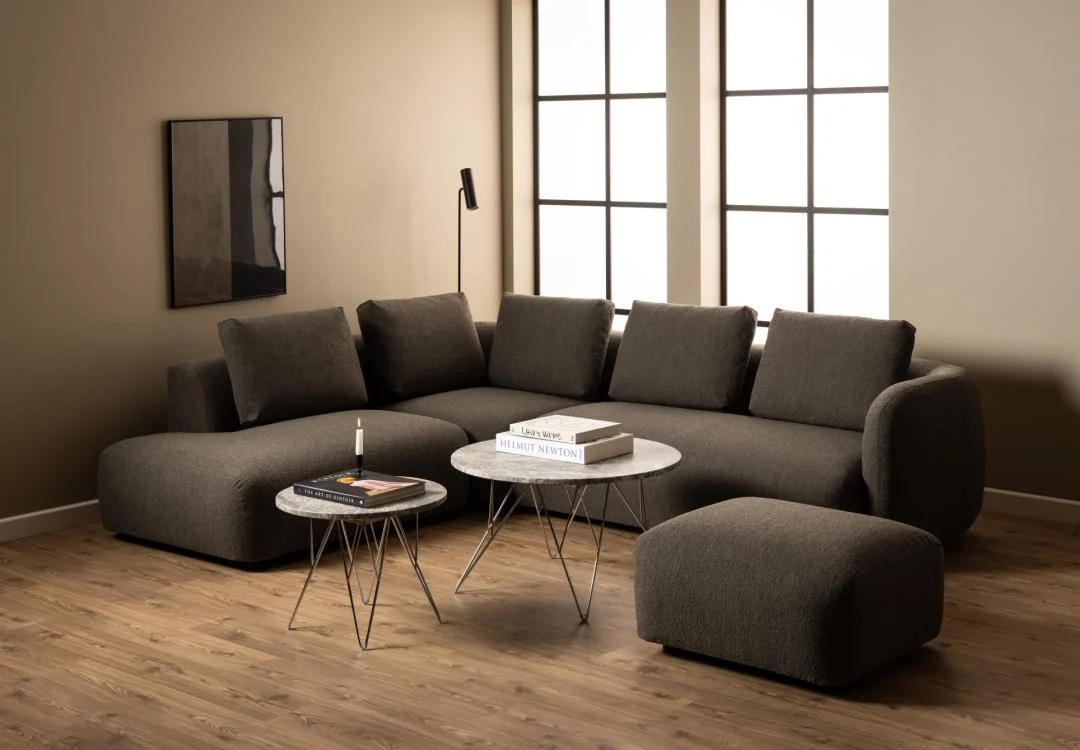 Een moderne woonkamer heeft een donkergrijze sectionele bank, twee 80 cm ronde grijsbruine Salontafels van kunststof, een boek en een kaars op de tafels, een houten vloer, beige muren, grote ramen en ingelijste abstracte kunst.