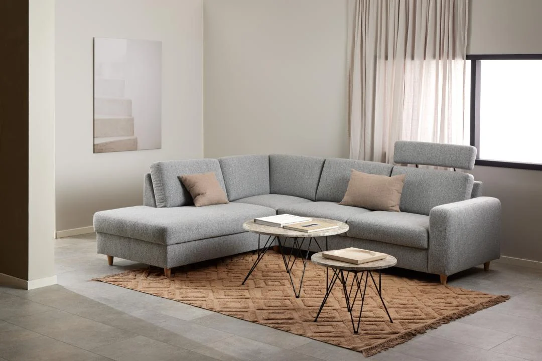 Een moderne woonkamer met een lichtgrijze L-vormige bank, twee beige kussens, twee ronde Bijzettafel Zwart, Grijs Rond Kunststof tafels, een vloerkleed met bruine structuur, beige gordijnen en een groot raam dat natuurlijk licht binnenlaat.