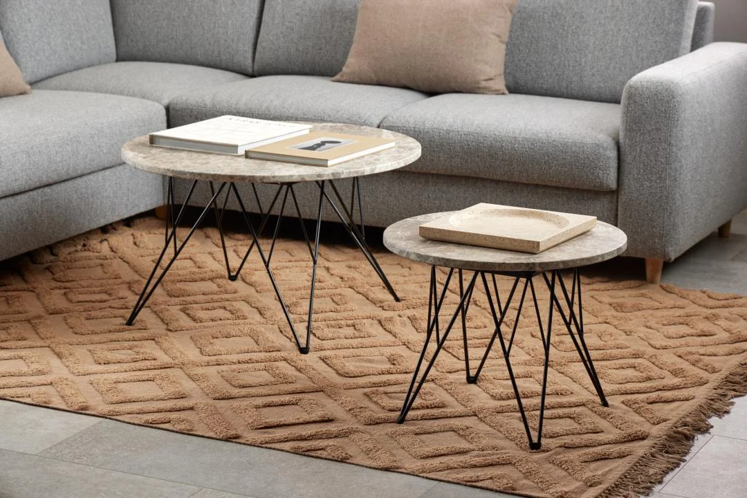 Een Bijzettafel Zwart, Grijs Rond Kunststof ligt op een bruin vloerkleed met structuur voor een grijze bank, met boeken en een dienblad er netjes bovenop.