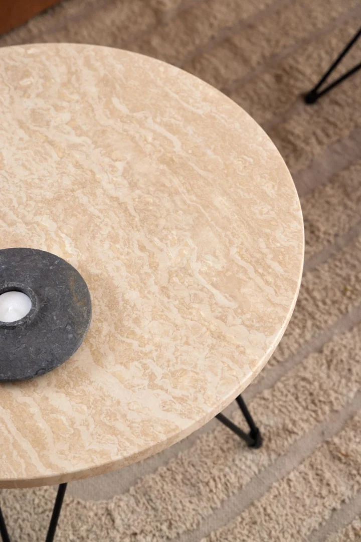 Een Bijzettafel Beige, Zwart 55 CM Rond van travertin met zwarte poten staat op een beige vloerkleed. Op de tafel staat een zwart stenen waxinelichthouder met een witte kaars.