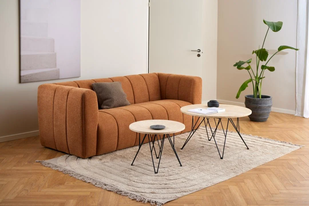 Een moderne woonkamer met een oranje bank, bruine kussens, beige vloerkleed, plant in pot, neutrale muren en twee Bijzettafel Beige, Zwart 55 CM Rond travertin salontafels. Een gesloten deur en muurkunst op de achtergrond.