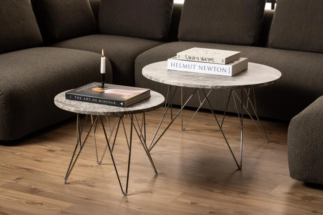 Een Bijzettafel Grijs, Bruin 55 CM Rond van kunststof staat op een houten vloer. Boeken en een kaars rusten op het oppervlak, terwijl een donkere sofa rond de tafel staat.